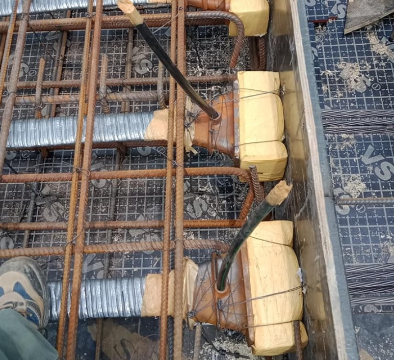 A detailed close-up photograph looking down at the edge formwork of a post tension slab. It shows multiple corrugated aluminum ducts, which will later hold multi-strand tendons, connecting to rectangular anchor heads that are heavily taped. Steel rebar grids and shear reinforcement cages frame the components.